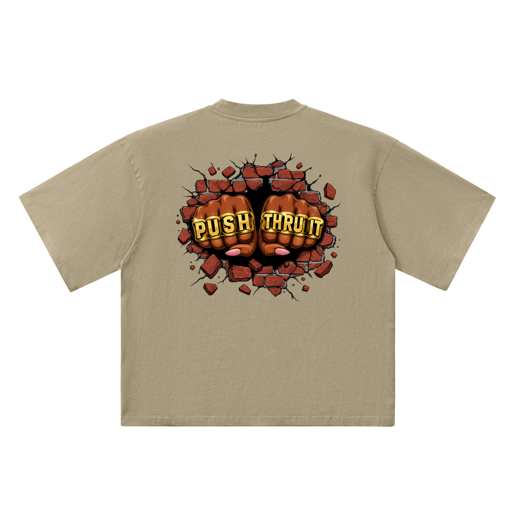 A beige short-sleeve t-shirt shown from the back, featuring a graphic of two clenched fists bursting through red bricks with the words 'PUSH THRU IT' in yellow letters.