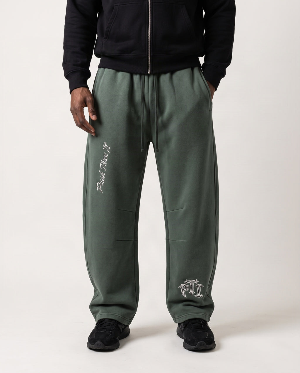 A man wearing olive green barrel leg sweatpants with metallic silver embroidered graphics, a black zip-up hoodie, and black sneakers, photographed against a plain light gray studio background.