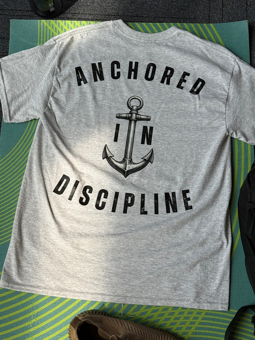 A heather gray t-shirt laid flat on a green and yellow striped blanket, showing the back with a black graphic of an anchor and the words 'ANCHORED IN DISCIPLINE'.