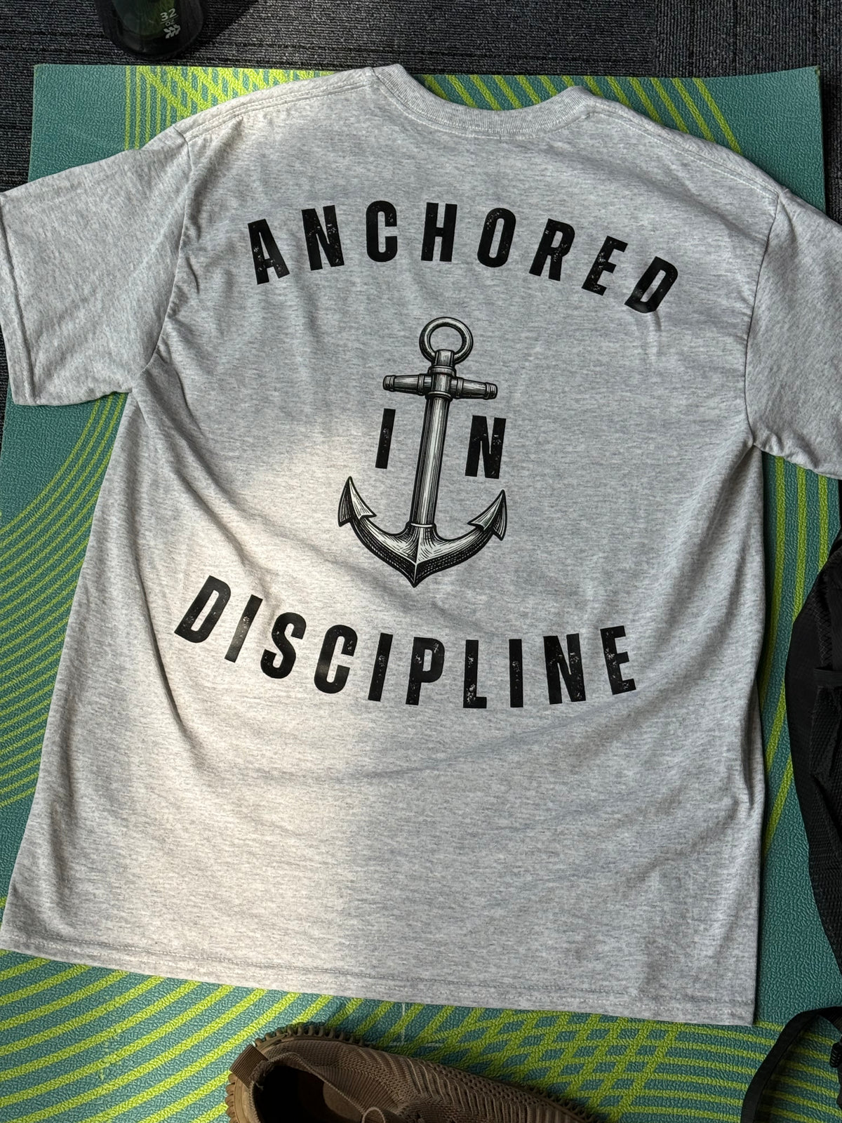 A heather gray t-shirt laid flat on a green and yellow striped blanket, showing the back with a black graphic of an anchor and the words 'ANCHORED IN DISCIPLINE'.