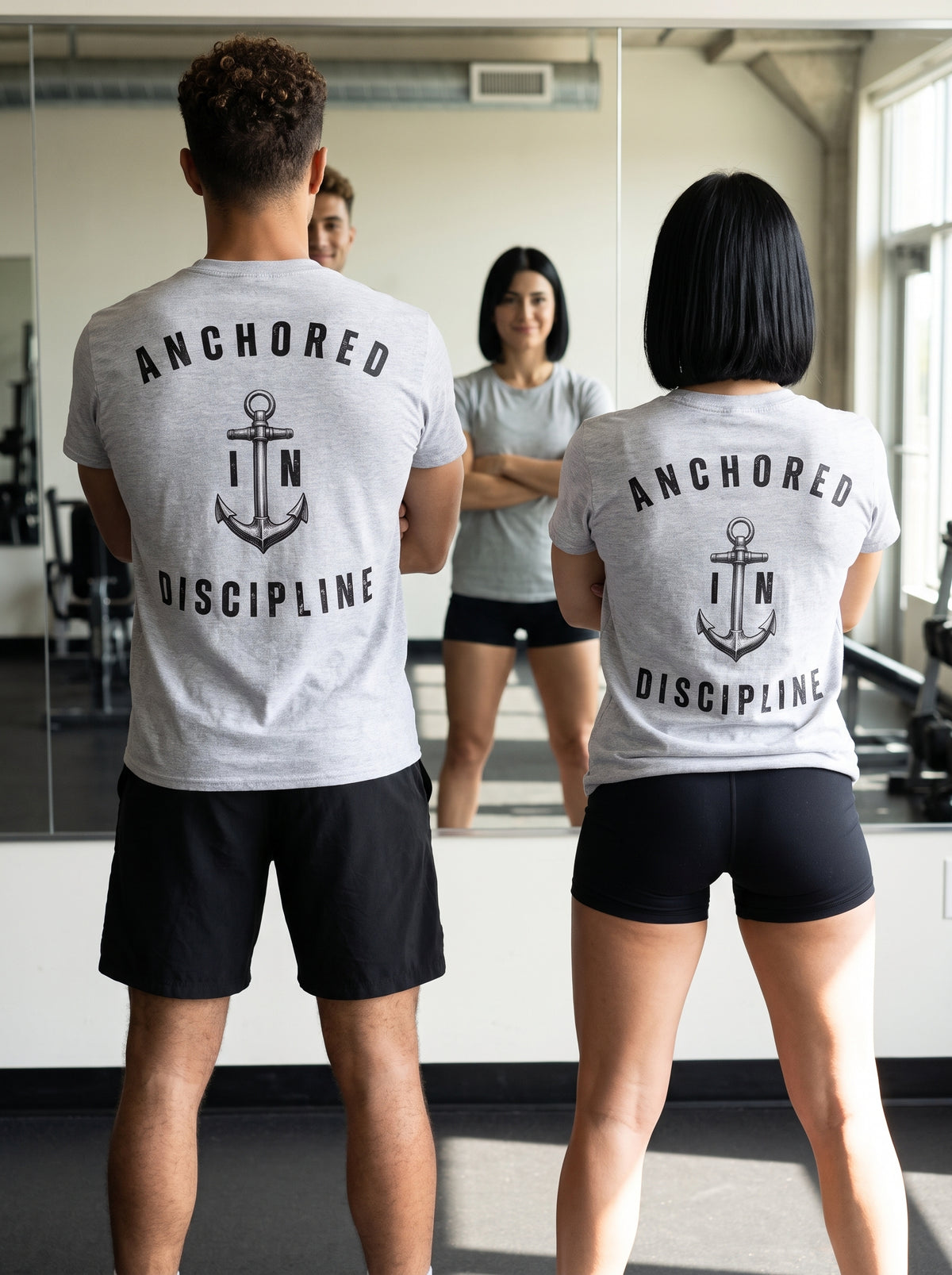 A man and a woman wearing matching light gray heather t-shirts with a black anchor graphic and the words 'ANCHORED IN DISCIPLINE' on the back, standing in front of a gym mirror.